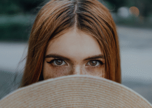 Frau hält Hut vors Gesicht und schaut mit intensivem Blick hervor - Symbol für Sichtbarkeit